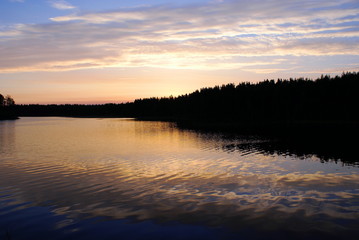 Obraz premium Early morning on the lake. On the horizon a dark forest. The sun has not risen yet, but has painted the clouds pink. Blue sky. The water in the lake is blue, the water reflects pink clouds.