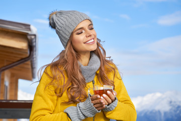 Woman drinking warm tea in the rustic wooden outdoor cafe on mountain, alpine view, snow on hills.