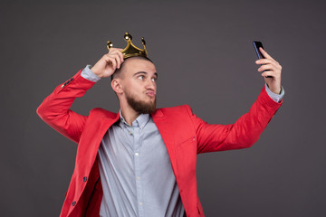 Young bearded handsome man in gold crown taking selfie looking at smartphone - obrazy, fototapety, plakaty