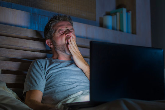 attractive tired and stressed workaholic man working late night exhausted on bed busy with laptop computer yawning feeling sleepy and overworked in project deadline stress