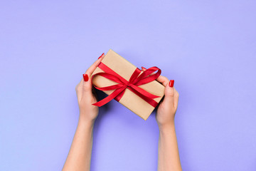 Woman hands give wrapped valentine or other holiday handmade present in paper with red ribbon. Present box, decoration of gift on purple table, top view with copy space