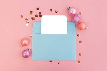 Frame of christmas balls and confetti with blank card mockup in blue envelope on pink background.