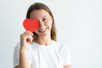 Playful young woman holding paper heart in front of her eye. Pretty girl looking at camera and smiling. Saint Valentines Day concept. Isolated front view on white background.