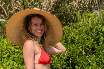 Young charming girl in a big straw hat stands among tropical palm trees in a swimsuit on a sunny summer day