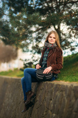 Fototapeta na wymiar Attrective young girl sits by the embankment. Blond hair and brown jacket. Spring