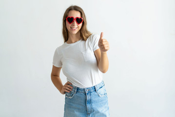 Woman wearing heart shaped sunglasses and showing thumb up. Smiling pretty young lady looking at camera. Recommendation concept. Isolated front view on white background.