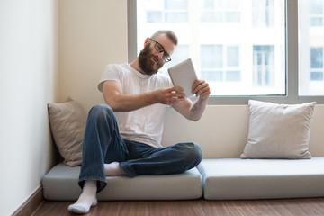 Content curious young man turning tablet while viewing picture. Concentrated bearded guy sitting on pillow. Leisure concept