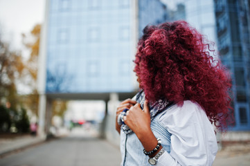 Attractive curly african american woman in jeans dress posed against modern multistory building.