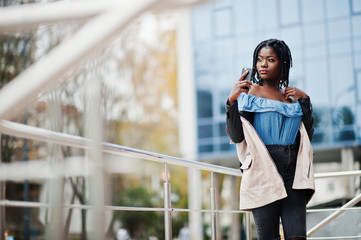 Attractive african american woman with dreads in jacket posed near railings against modern multistory building making selfie at mobile phone.