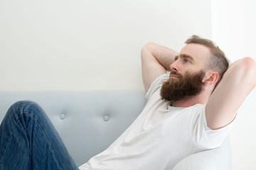 Sad fired employee depressed about job loss. Bearded young man in casual resting on couch at home and staring into vacancy. Unemployment or depression concept