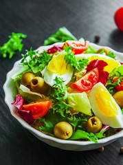 various fresh mix salad leaves with tomato, eggs and olive in bowl on dark wooden background