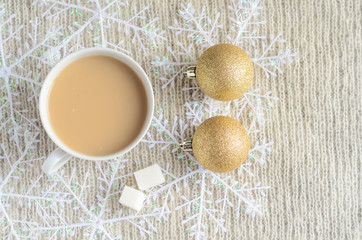 A Cup of coffee with milk, two pieces of sugar ,decorated snowflakes and two pair Christmas golden balls on a light wool sweater.Winter concept.