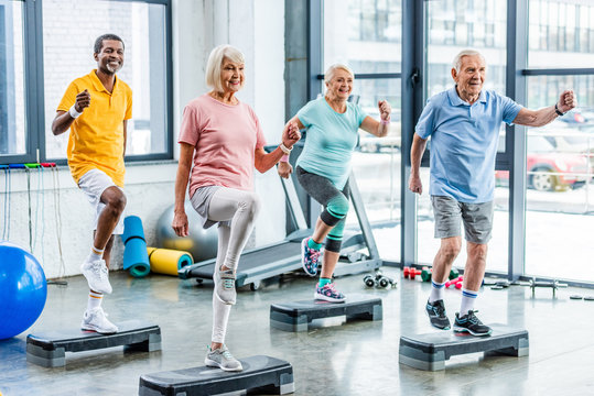 smiling multiethnic senior athletes synchronous exercising on step platforms at gym