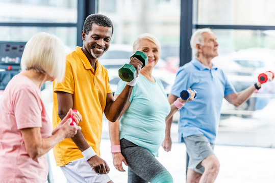 laughing african american man and his friends making exercise with dumbbells at gym