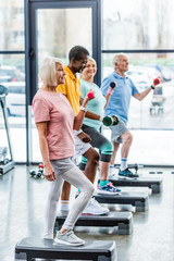 side view of senior multicultural sportspeople synchronous exercising with dumbbells on step...
