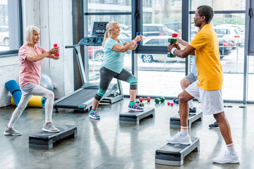 selective focus of senior multicultural sportspeople synchronous exercising with dumbbells on step...