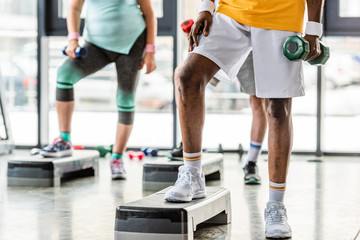 cropped image of african american sportsman and his friends exercising with dumbbells on step platforms at gym