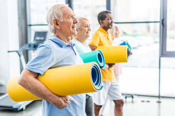 Fototapeta premium side view of multethnic senior athletes holding fitness mats at sports hall