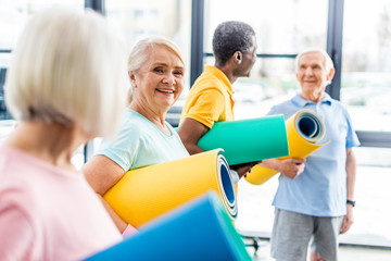 Obraz premium selective focus of senior sportswoman holding fitness mat and her friends standing behind at gym