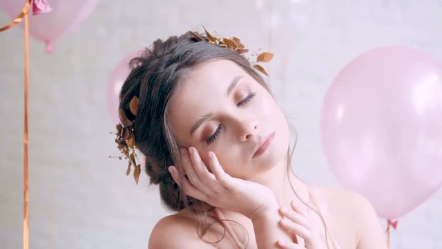 attractive brunette girl with cute baby face poses as model, charming drowsy angel with soft features and gentle hairstyle with golden wreath on her head, bare shoulders, pink balloons on background