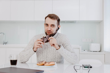 A man with a beard is sitting at the table in the white kitchen and eating a chocolate donut for lunch
