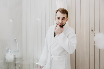 A man with a beard in a white bathrobe  is going to go into the shower