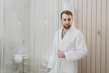 A man with a beard in a white bathrobe  is going to go into the shower