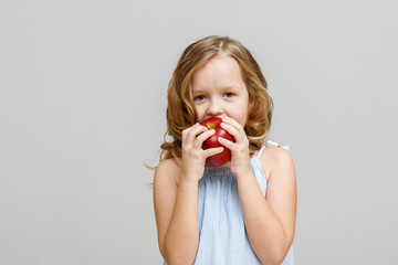 Portrait of a happy smiling little blonde girl on a gray background. Baby eating red apple