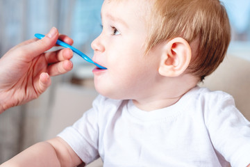 Mom feeding the baby holding hand with a spoon of food in the kitchen. Healthy baby nutrition. Emotions of a child