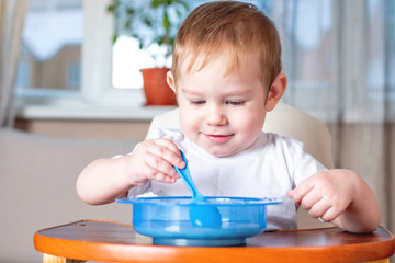 Little cute baby learning to eat with a spoon himself at the kids table in the kitchen. Healthy baby food