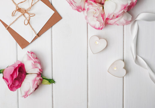 Flowers, heart shaped candles and paper envelopes on the white wooden background.