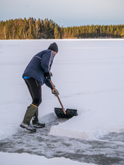 Man is preparing snow for ice hockey rink outdoor with a shovel on the frozen lake.