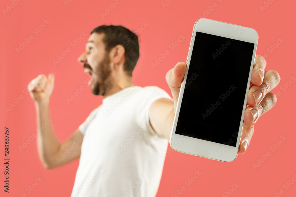 Wall mural Young handsome man showing smartphone screen over coral background with a surprise face. Human emotions, facial expression concept. Trendy colors