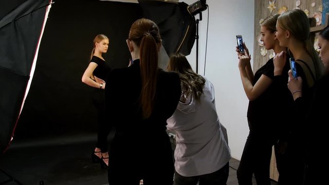 Young woman model having a photo session in the studio. Shooting the model in black clothes. Shooting in full height. Another models shooting the backstage on their phones