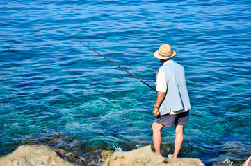 A man with a fishing rod on a sea stony shore catches fish. View from the back. Cyprus, Paphos, waterfront Coral Beach Hotel & Resort Cyprus in June 2017.