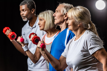 side view of senior mutliethnic sportspeople synchronous exercising with dumbbells on black with spotlight