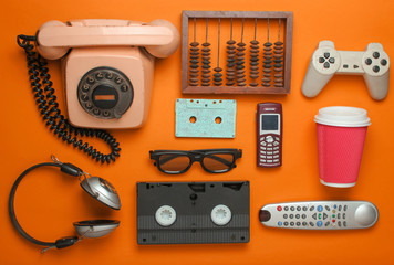Retro objects on orange background. Rotary telephone, audio cassette, video cassette, gamepad, 3d glasses, tv remote, headphones, push-button phone. Analog media technology of the past. Flat lay.. - obrazy, fototapety, plakaty