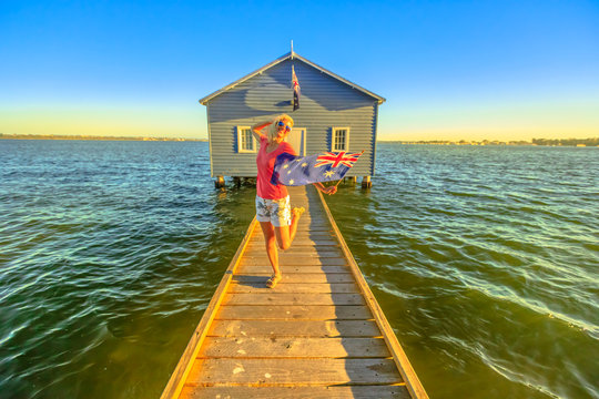 Happy caucasian woman with australian flag enjoying in front of Blue Boat House with wooden jetty on Swan River at sunset. Perth landmarks in Western Australia. Copy space.