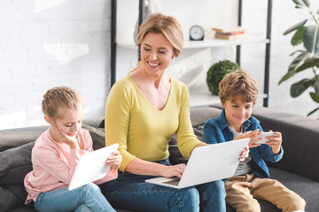 happy mother looking at cute little kids using digital devices at home