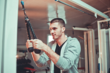 Sporty man in hoody exercising triceps muscles in the gym with pull-down ropes fitness equipment machine attachments. Training process.