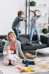 exhausted mother holding infant child and looking at camera while naughty kids playing at home