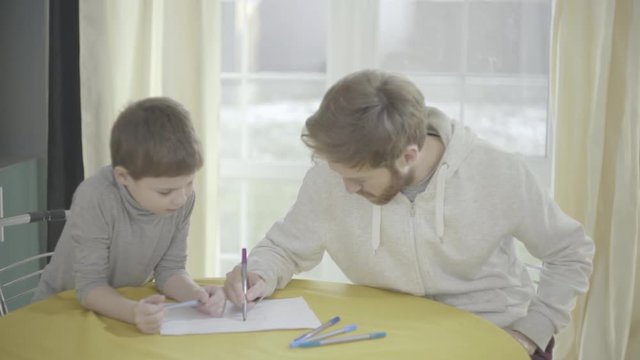 Bearded father and his little son paint on paper sitting in living room at the table. Father-child relationship.