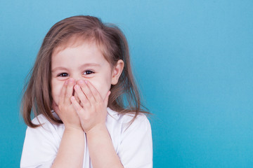 Little girl on a blue background laughs covering her face with her hands