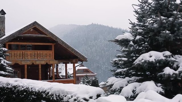 Christmas wooden mansion in mountains on snowfall winter day. Cozy chalet on ski resort near pine forest. Cottage of round timber with wooden balcony. Fir-trees covered with snow. Chimneys of stone.