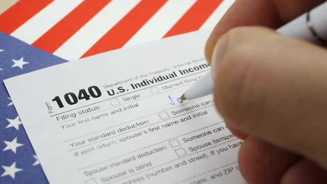Hand filling american 1040 Individual Income Tax return form on table with US flag at home