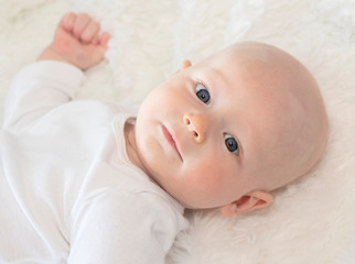 Beautiful 6 Month Baby Boy Dressed in White & Lying on Fluffly White Blanket Looking at Camera. Smiling & Happy