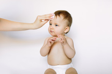 mother hand cleans face of a one year baby girl whith a cotton pad on white background. Baby hygiene, care, healthcare, medical concept.