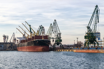 Fototapeta premium Large cargo ship in the seaport during loading. Cranes load the cargo into the ship. The work of the seaport. Industrial seascape on a daytime