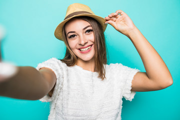 Young cheerful attractive woman in straw hat taking selfie on the camera of her phone on the blue background.