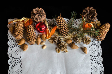 The top half of an openwork paper napkin with various cones, a fir branch, old worn Christmas toys,almonds and orange peel on a black background

